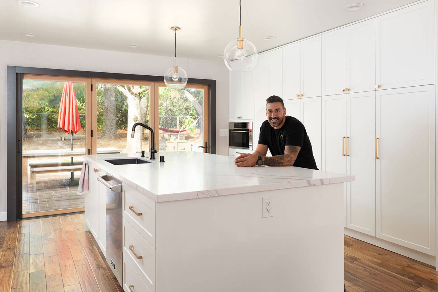 The image shows a man standing in a modern kitchen with white cabinets, a center island with a sink, and stainless steel appliances, including a refrigerator and oven. He is wearing a black shirt and has his hands on the countertop. There s a dining table visible through an open doorway to another room. The flooring appears to be hardwood, and there are various kitchen accessories such as a red napkin dispenser, a knife block, and some decorative items on the countertops. The kitchen has a warm ambiance with natural light coming from windows not visible in the image.