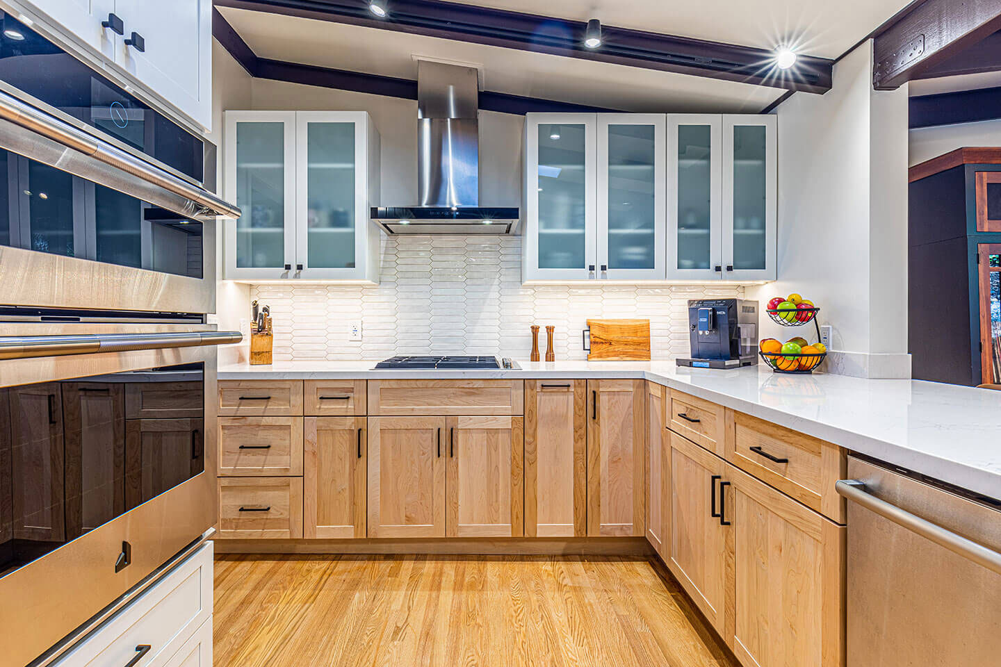 This is a photograph of a modern kitchen interior featuring light-toned wooden cabinets, stainless steel appliances, granite countertops, white subway tile backsplash, and hardwood flooring.