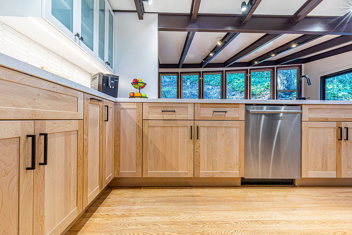The image shows a modern kitchen with light-colored wooden cabinets, stainless steel appliances, including a refrigerator, oven, and dishwasher, a sink, and a hardwood floor.