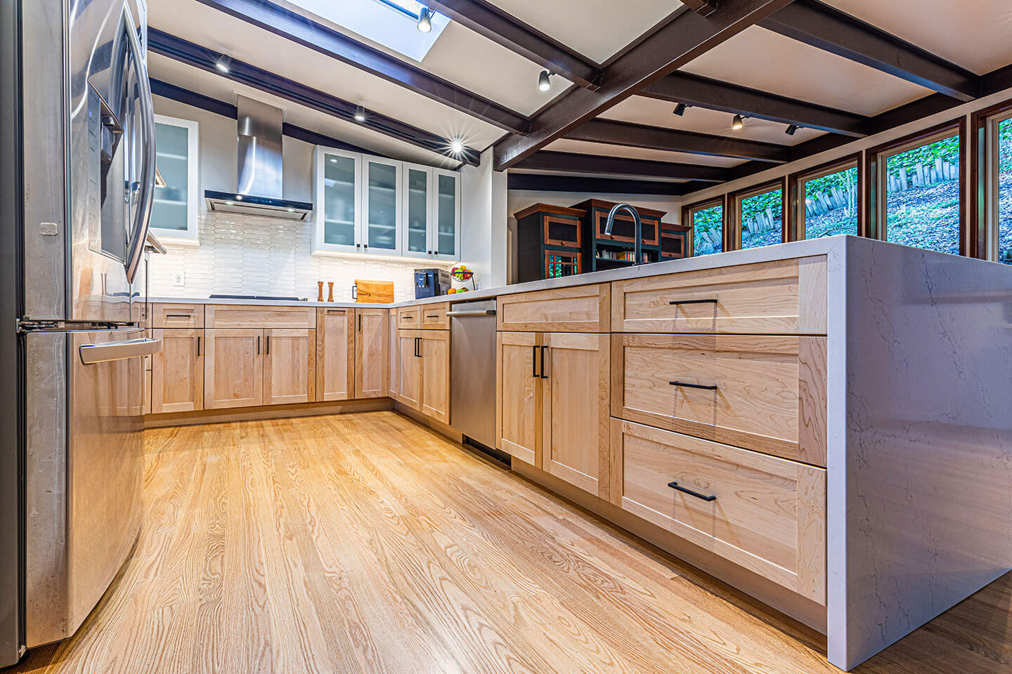 The image shows a modern kitchen interior with light-colored wood cabinets, stainless steel appliances, hardwood flooring, and a large island in the center.
