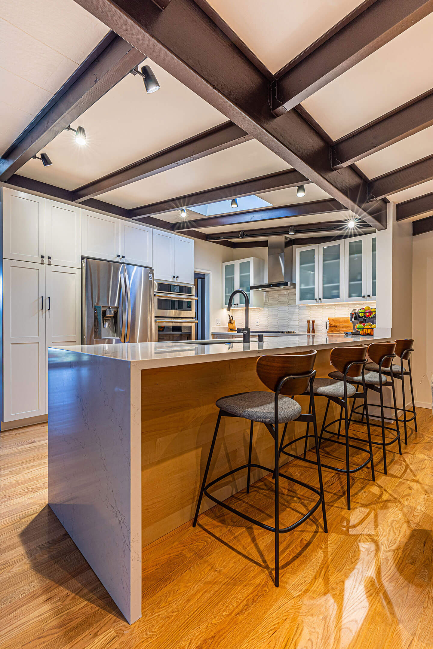 The image shows a modern kitchen interior with a center island, bar stools, wooden cabinets, stainless steel appliances, and recessed lighting.