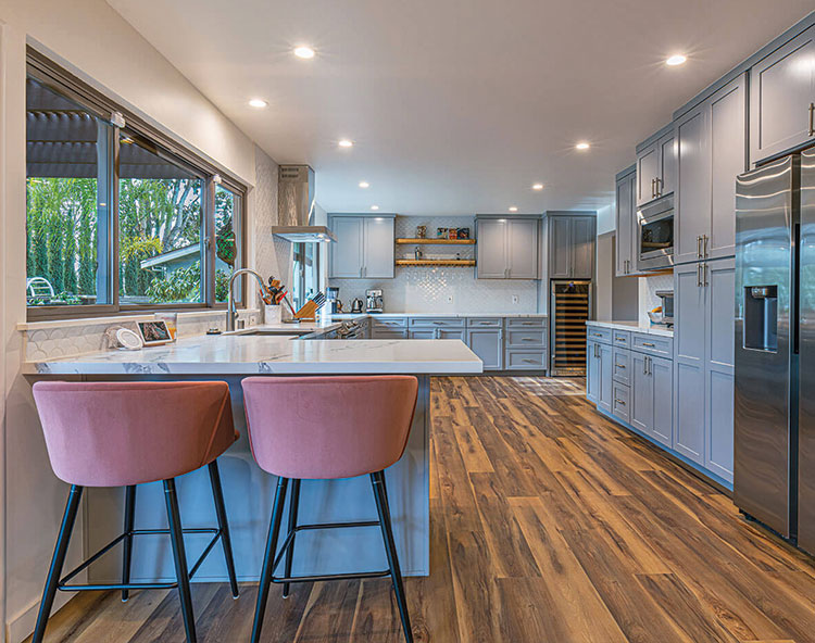 The image shows a modern kitchen interior with a sleek design, featuring light-colored cabinets, a large island with barstools, a hardwood floor, and stainless steel appliances.