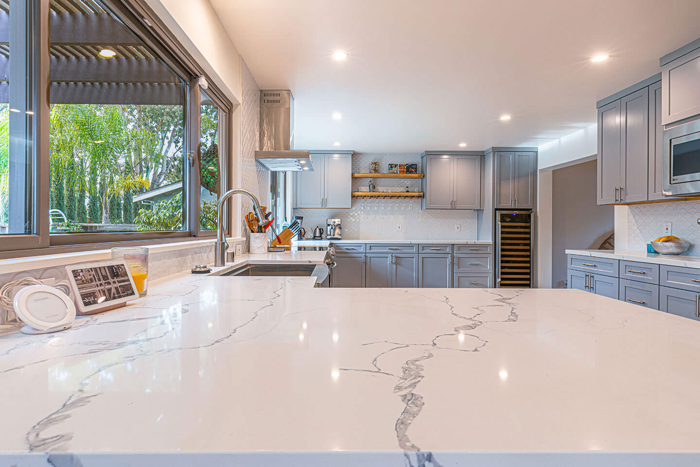 A modern kitchen with a large island featuring marble countertops and a backsplash, stainless steel appliances, white cabinets, and a window overlooking a patio area.