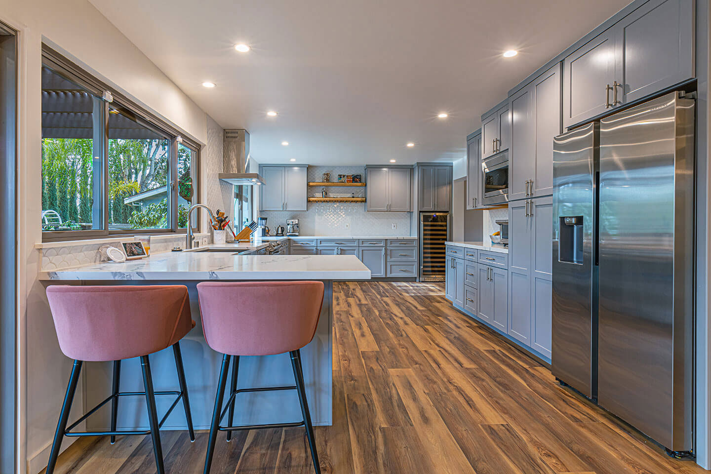 The image shows a modern kitchen interior with stainless steel appliances, including a refrigerator, oven, and microwave, dark wood cabinets, a central island with bar stools, a countertop with a sink, and a hardwood floor.
