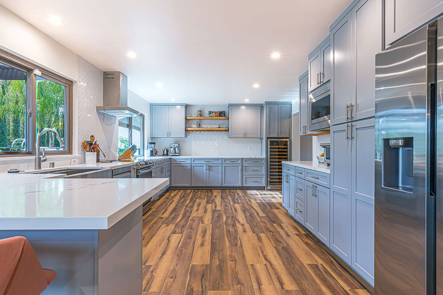 The image shows a modern kitchen interior with a large island countertop, high-gloss cabinets, stainless steel appliances, wood flooring, and a clean, organized appearance.