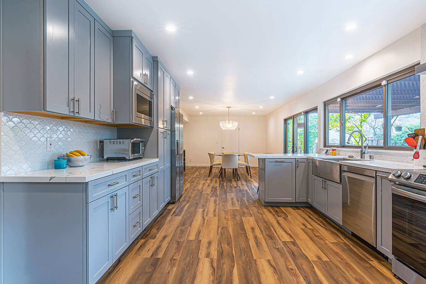 The image shows a modern kitchen interior with light wood flooring, gray cabinets, stainless steel appliances, and a large central island with a sink.