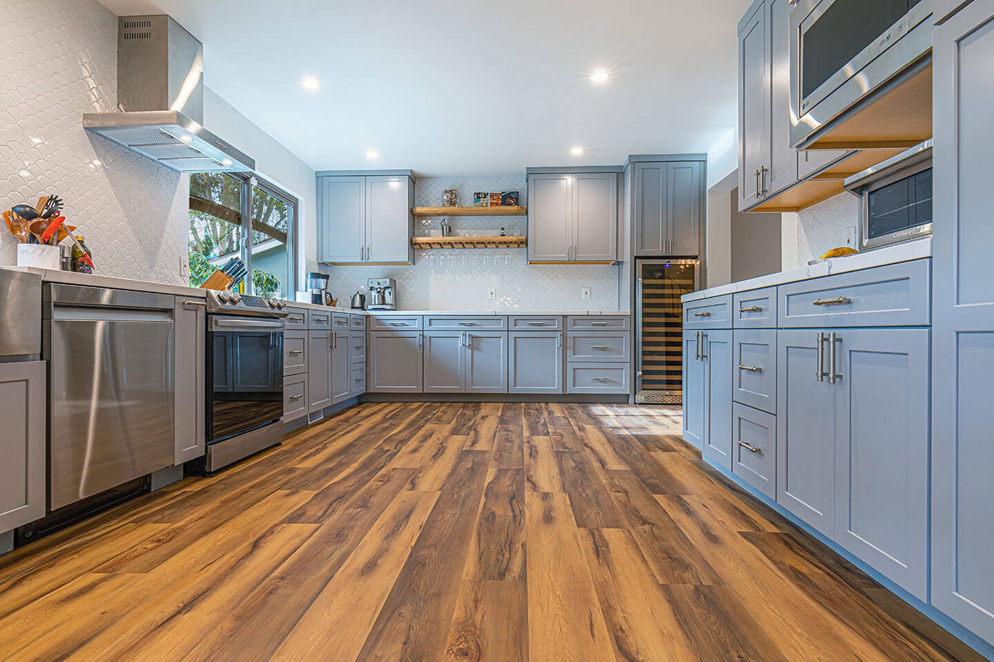 The image shows a modern kitchen with dark wooden flooring, sleek stainless steel appliances including a refrigerator, oven, and microwave, and a variety of cabinetry in shades of gray and blue.
