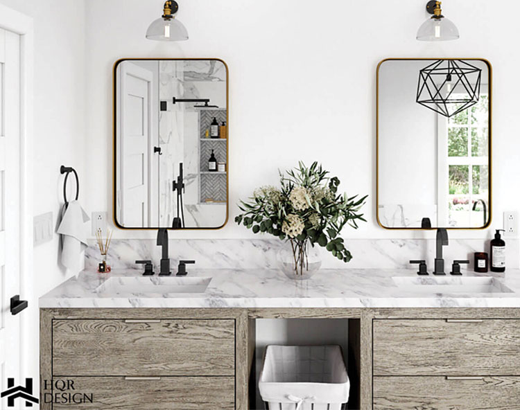 The image shows a modern bathroom interior with a double vanity featuring marble countertops, gold-framed mirrors, and a white sink. A trash can is tucked under the counter, and the wall behind the vanity has a geometric pattern. Above the vanity, there are two framed mirror panels, and decorative items are placed on the countertop. The room is well-lit with natural light coming from a window to the right.