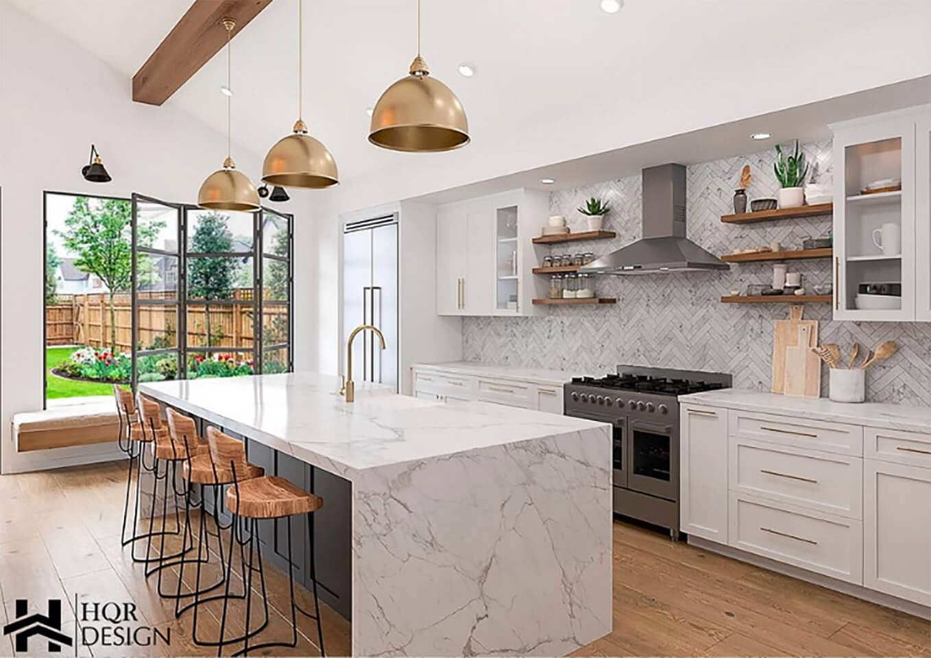The image displays a modern kitchen interior with marble countertops, white cabinets, a central island with barstools, pendant lighting, a large window allowing natural light to enter, and various kitchen appliances such as an oven and refrigerator.