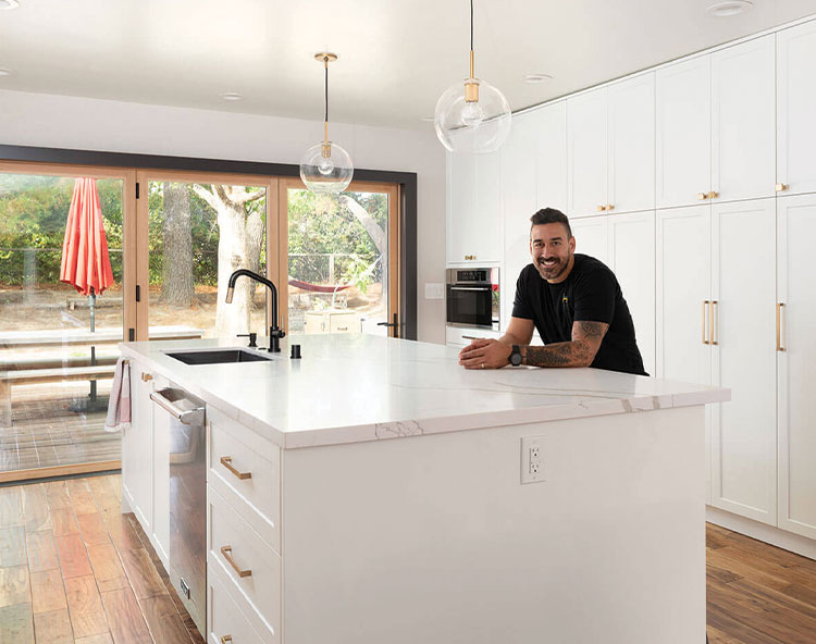 A man is standing at a kitchen island with a white countertop, wearing a black t-shirt, and looking directly at the camera. The kitchen has white cabinets, stainless steel appliances, and a modern design.