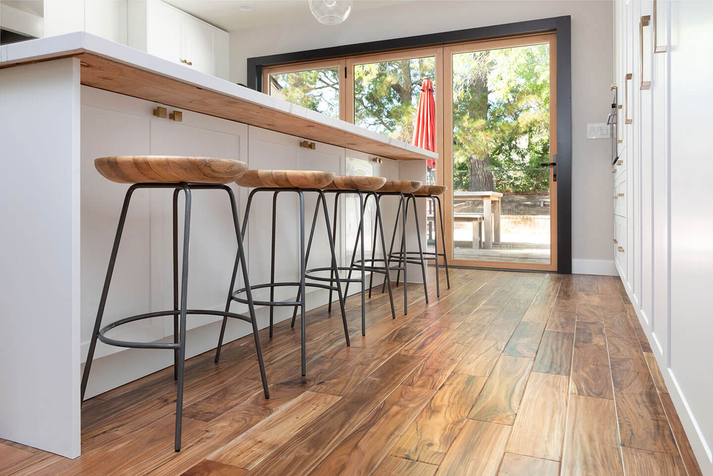 A modern kitchen with dark hardwood flooring, black metal bar stools, a wooden countertop, and a white cabinet with a glass front.