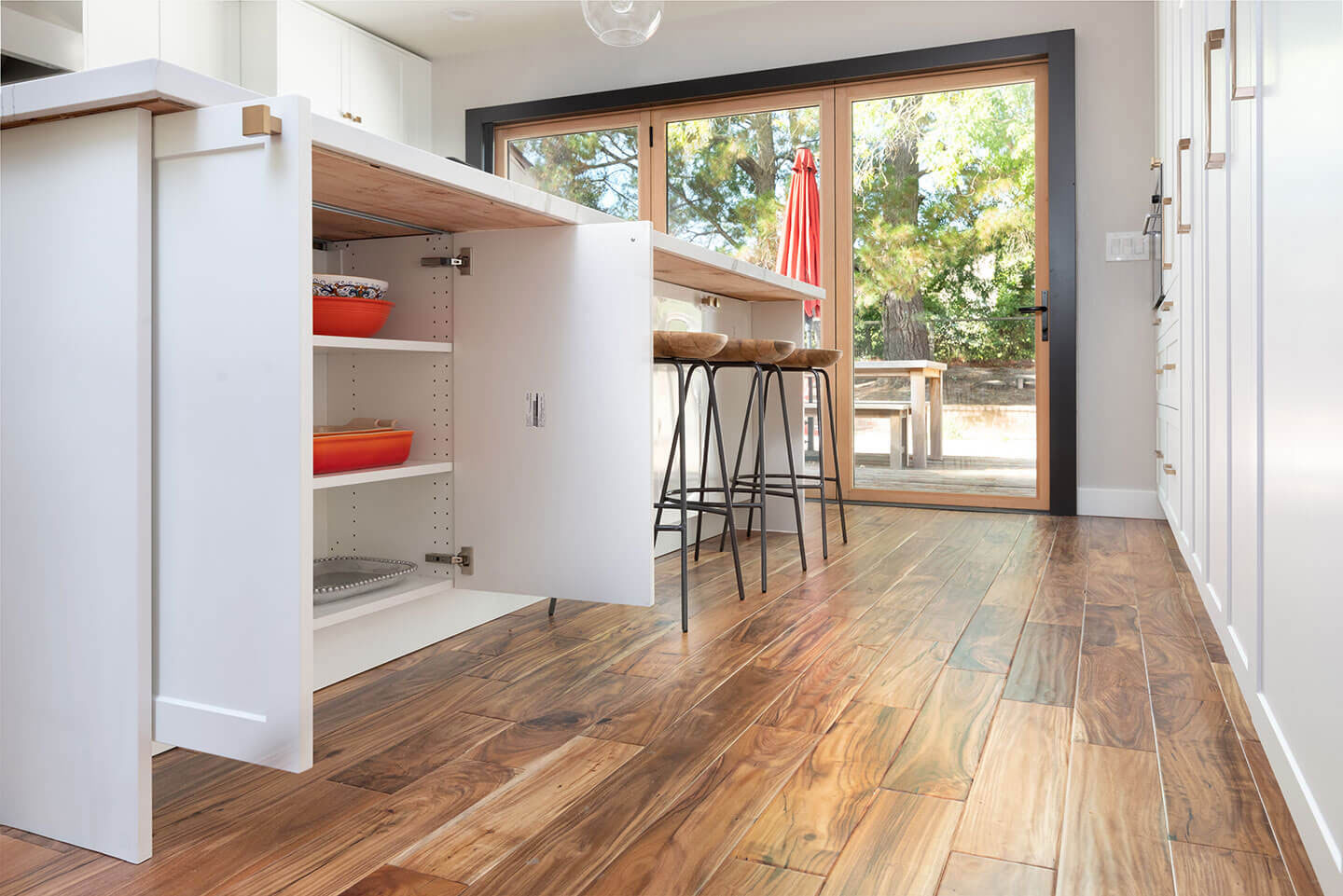The image shows a well-lit kitchen interior with hardwood flooring and modern cabinetry, featuring an island with bar stools, a built-in wine rack, and a clean, organized appearance.