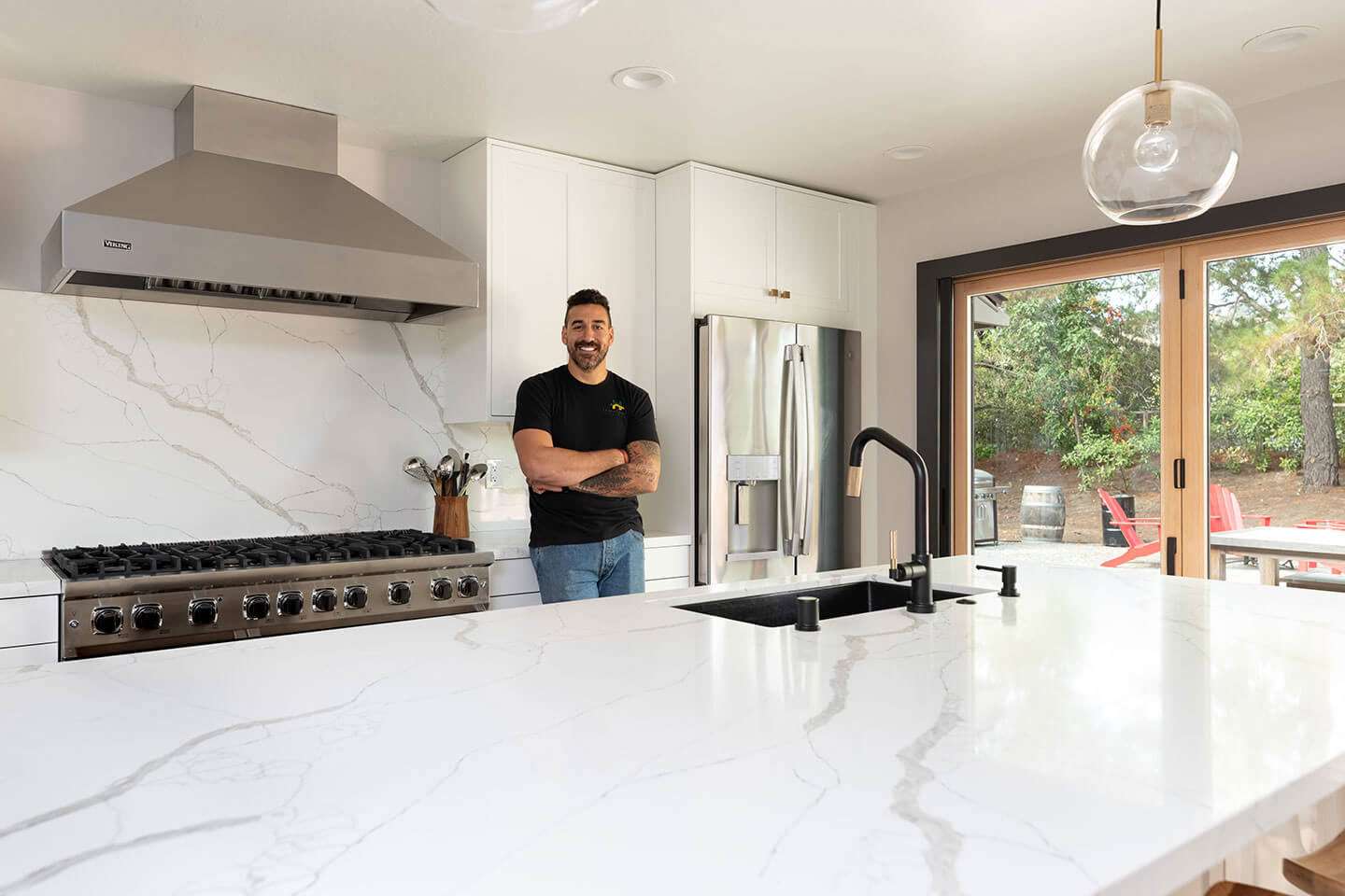 A man stands in front of a large kitchen island with marble countertops and stainless steel appliances, including a refrigerator and oven, in a modern home setting.
