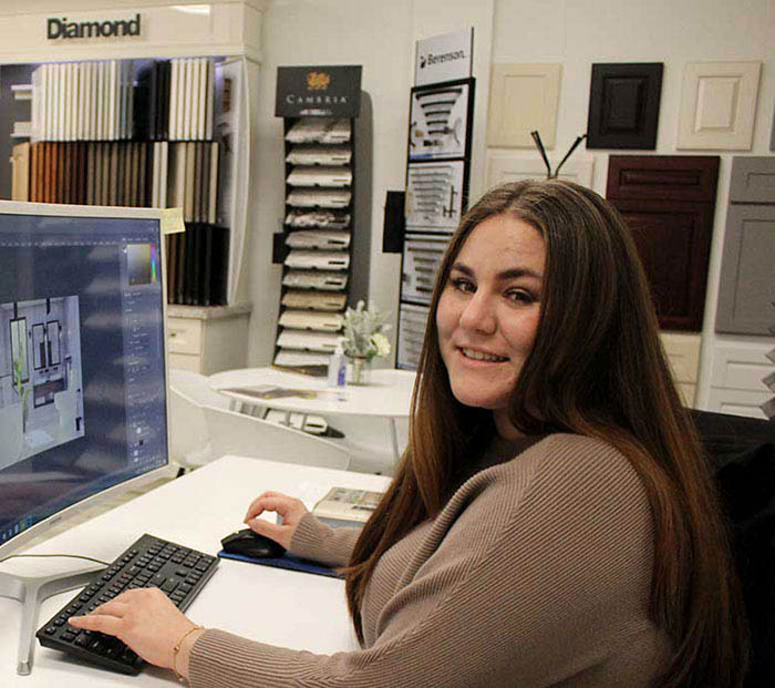 The image shows a woman sitting at a desk with a computer monitor displaying a design layout, working in an office setting with a variety of materials displayed on shelves behind her.
