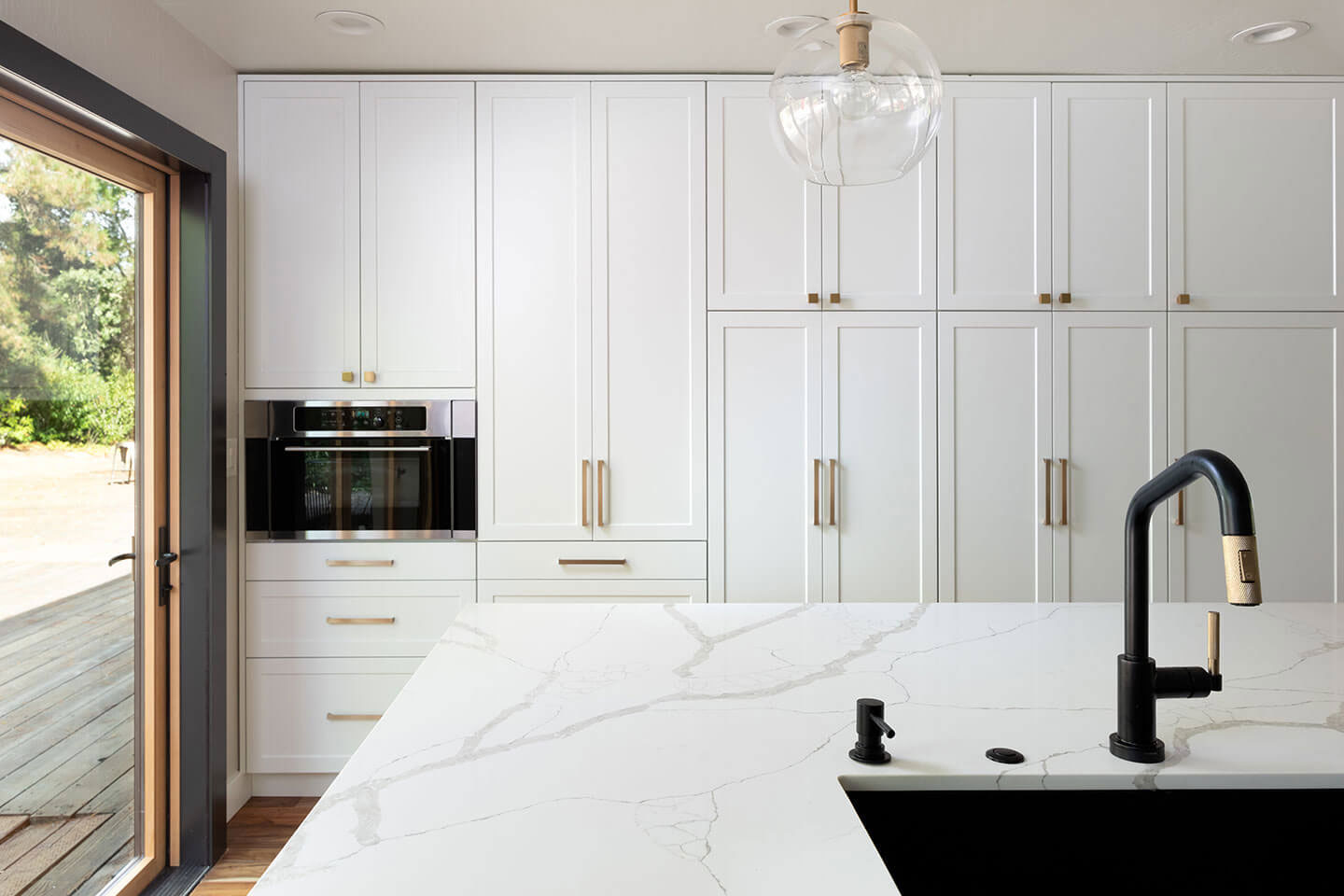 The image shows a modern kitchen interior with white cabinetry, marble countertops, stainless steel appliances, a black faucet, and a large island with a sink in the center.