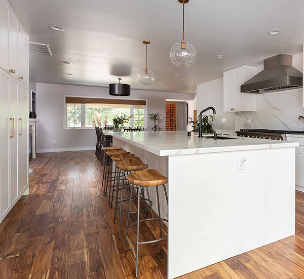 The image shows a modern kitchen interior with a large island countertop, bar stools, wooden flooring, white cabinets, stainless steel appliances, and a contemporary design aesthetic.